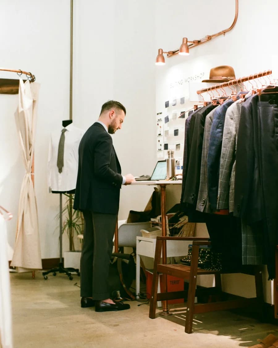 A tailor standing at a laptop on a bench in his own suiting workshop, racks of jackets beside him.