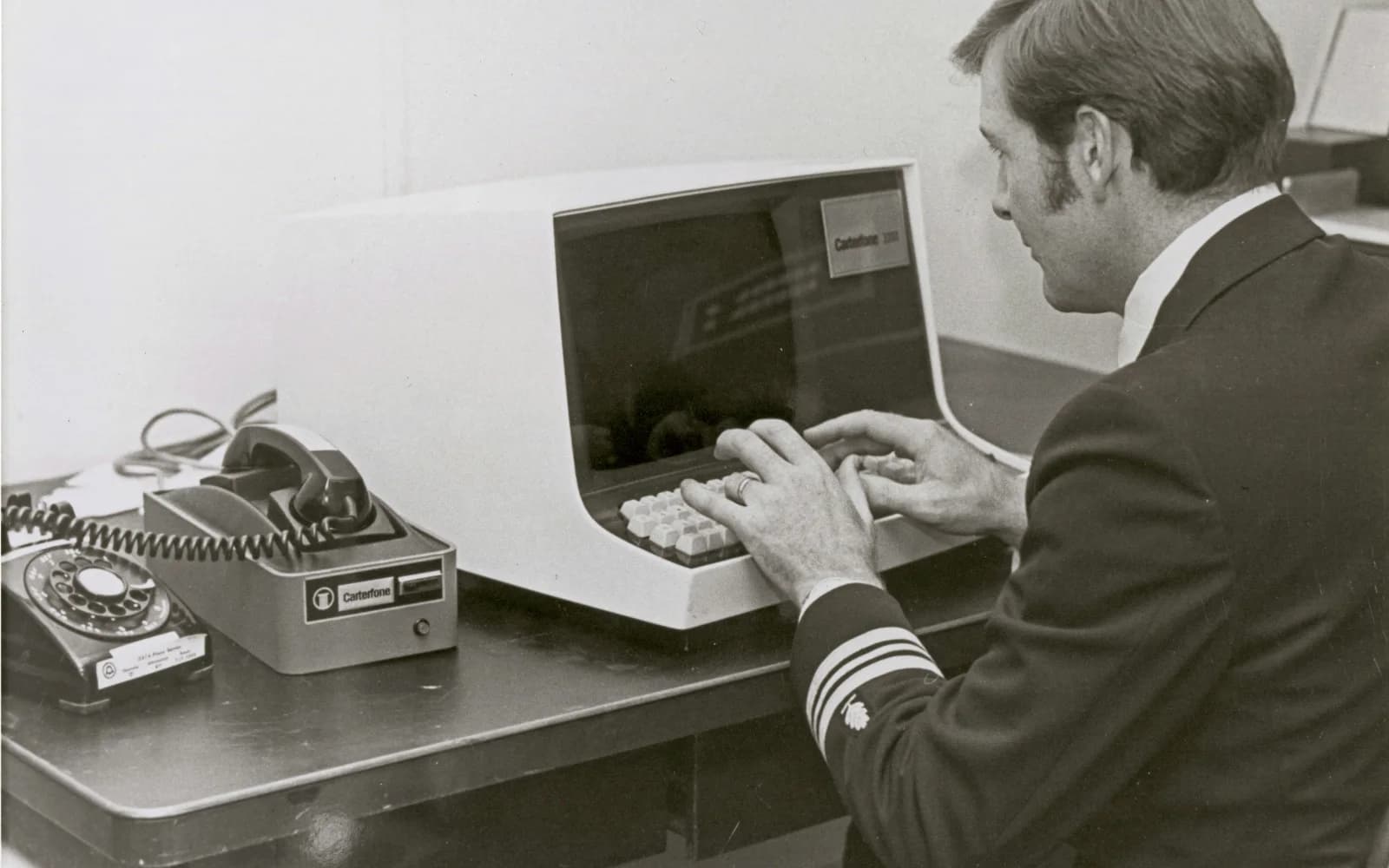 An officer in the 1970s typing at a computer terminal, a rotary telephone on the desk beside him.