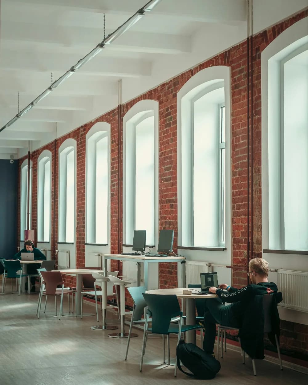 Two people working alone at desks under tall arched windows in a converted brick hall.